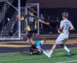 High school soccer match featuring player 16 jumping over a diving goalkeeper while player 2 watches. Nighttime action shot.
