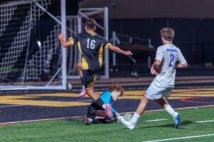 High school soccer match featuring player 16 jumping over a diving goalkeeper while player 2 watches. Nighttime action shot.