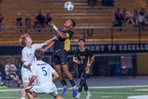 High school soccer match: players compete for the ball in mid-air under stadium lights, capturing an intense moment.
