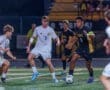 High school soccer players compete under floodlights; one dribbles the ball while others defend.