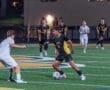 High school soccer players compete on a lit field, with one dribbling the ball while defenders advance towards him.