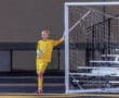 Young soccer goalkeeper in a bright yellow uniform, leaning against the goalpost during a night game.