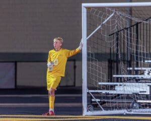 Young soccer goalkeeper in a bright yellow uniform, leaning against the goalpost during a night game.