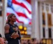 Athlete with braided hair speaks passionately, hand on heart, in front of an American flag background during a sports event.