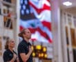 Coaches stand with hands over hearts during the national anthem, with a vibrant American flag displayed in the background.
