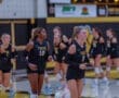 Two volleyball players jog on the court, wearing black jerseys with Tigers as teammates prepare for a game.