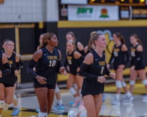 Two volleyball players jog on the court, wearing black jerseys with Tigers as teammates prepare for a game.