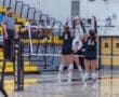 High school volleyball match action featuring players blocking a spike at the net, with a referee observing.