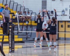 High school volleyball match action featuring players blocking a spike at the net, with a referee observing.