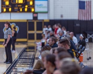 Spectators watch a basketball game with a referee in focus and an American flag in the background.