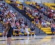 Young volleyball player serving the ball during a match in a crowded gym with yellow and black seating.