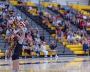 Young volleyball player serving the ball during a match in a crowded gym with yellow and black seating.