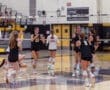 Girls' volleyball team celebrates a point during a match in a gym, showcasing teamwork and excitement.