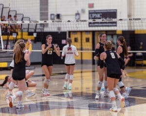 Girls' volleyball team celebrates a point during a match in a gym, showcasing teamwork and excitement.