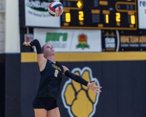 Volleyball player serves the ball during a game, displaying focused determination against the scoreboard backdrop.