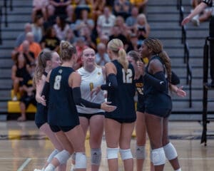 Volleyball team in a huddle celebrating teamwork during a match, with excited fans in the background.