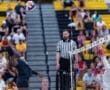 A volleyball player spikes the ball over the net during a competitive match with a cheering crowd in the background.