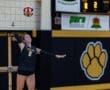Volleyball player in a Tigers uniform serves the ball during a match, with scoreboard in the background.