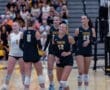 Five female volleyball players in team uniforms celebrate during a game, showcasing teamwork and excitement.