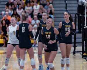 Five female volleyball players in team uniforms celebrate during a game, showcasing teamwork and excitement.