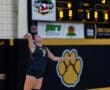 Female volleyball player prepares to serve during a match, showcasing athleticism in a gym with vibrant team colors.