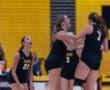 Cheering volleyball team in black jerseys celebrating a point, with excitement and teamwork showcased in a vibrant gym.