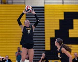 Volleyball player in black jersey sets the ball during a match, with teammates and spectators in the background.