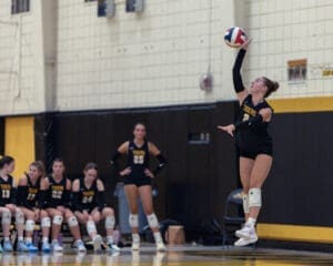 Volleyball player serving the ball during a game, with teammates observing on the bench in the background.