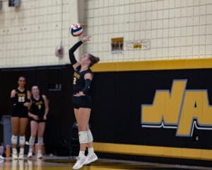 Female volleyball player from the Tigers team serves the ball during a match, with teammates in the background.