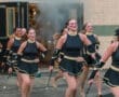 Marching performers in sparkling black costumes with twirling batons, smiling as they parade past a smoky entrance.