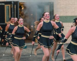 Marching performers in sparkling black costumes with twirling batons, smiling as they parade past a smoky entrance.