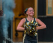 Cheerleader performing with pom-poms, smiling, in a lively outdoor setting during a sports event.