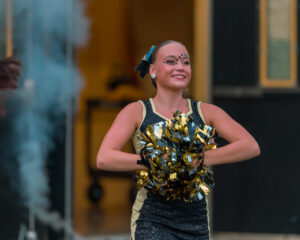 Cheerleader performing with pom-poms, smiling, in a lively outdoor setting during a sports event.