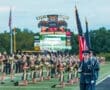 Color guard presents flags in front of a cheering marching band at a vibrant football game on a school field.