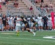 A football player in black tackles an opponent in white on a grassy field during a game with spectators in the background.