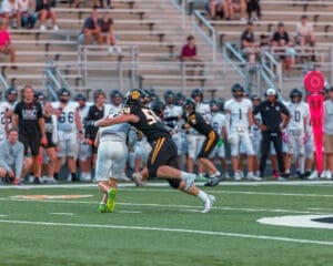 A football player in black tackles an opponent in white on a grassy field during a game with spectators in the background.