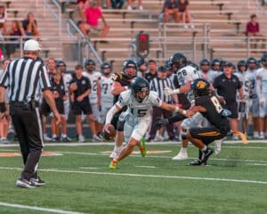 Quarterback evades a tackle during a high school football game, with players and spectators in the background.