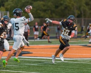Quarterback #15 throws a pass under pressure from a defender in a high school football game on a turf field.
