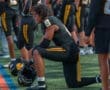 Football player in black uniform kneeling on the field, preparing for the game with teammates in the background.