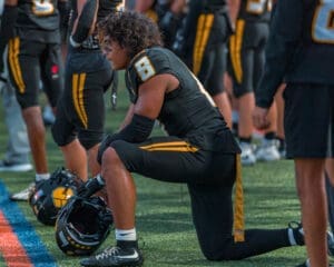 Football player in black uniform kneeling on the field, preparing for the game with teammates in the background.