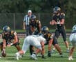 High school football players in black jerseys set for a play on the field during a game with a referee in the background.