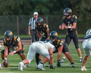 High school football players in black jerseys set for a play on the field during a game with a referee in the background.