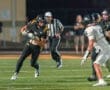 High school football player in black uniform dodges an opponent on the field during a night game under bright lights.