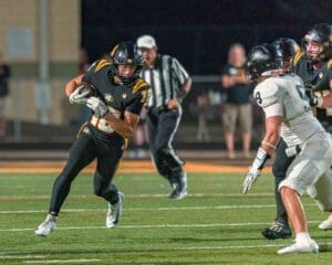 High school football player in black uniform dodges an opponent on the field during a night game under bright lights.