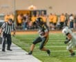 Football player in black uniform dodges defender while running on the field during a nighttime game. Referee watches closely.