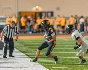 Football player in black uniform dodges defender while running on the field during a nighttime game. Referee watches closely.