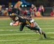 High school football player dives to catch the ball while being tackled, showcasing intense athletic action on the field.