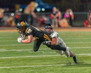 High school football player dives to catch the ball while being tackled, showcasing intense athletic action on the field.