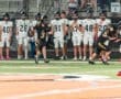 High school football game action, with a player in black running with the ball while teammates observe from the sidelines.