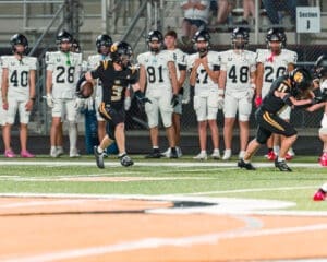High school football game action, with a player in black running with the ball while teammates observe from the sidelines.